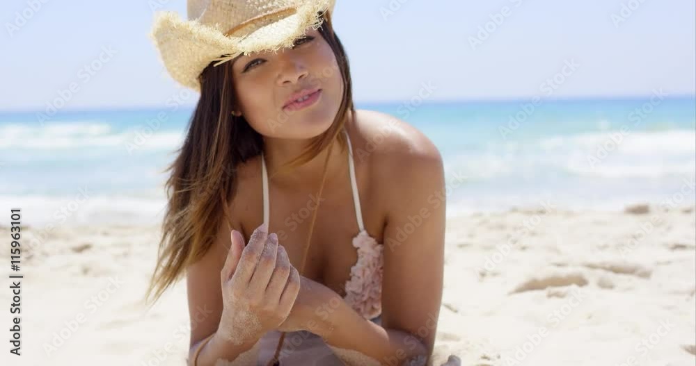 Laughing woman in hat and bikini holding something while on the beach with ocean waves in the background Laughing woman in hat and bikini holding something while on the beach with ocean waves in the background