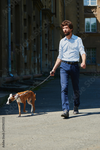 A man in a shirt and a beard, walks with a dog on a leash in the city