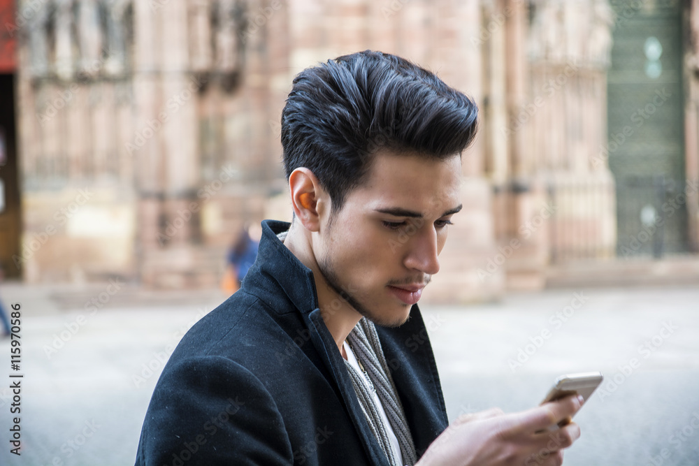 Young dark haired man standing in European city, using smartphone to send text message or surfing the internet