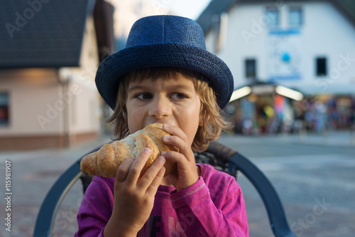 Cute little boy with blue hat sitting at the table eating tasty croissant.