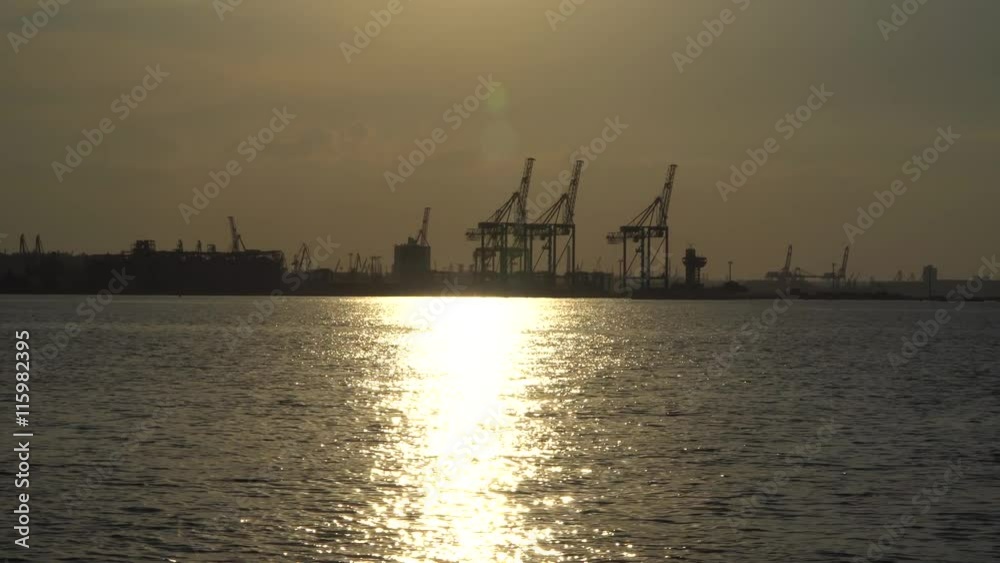 View of the sea port with the ship sailing at sunset. Visible cranes port, reflection of the sun. Active camera View of the sea port with the ship sailing at sunset. Visible cranes port, reflection of the sun. Active camera