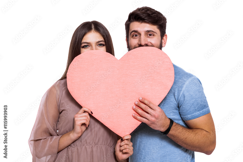Love is a great feeling! Beautiful young loving couple peeping out of big pink paper heart isolated on white background. Love is a great feeling! Beautiful young loving couple peeping out of big pink paper heart isolated on white background.