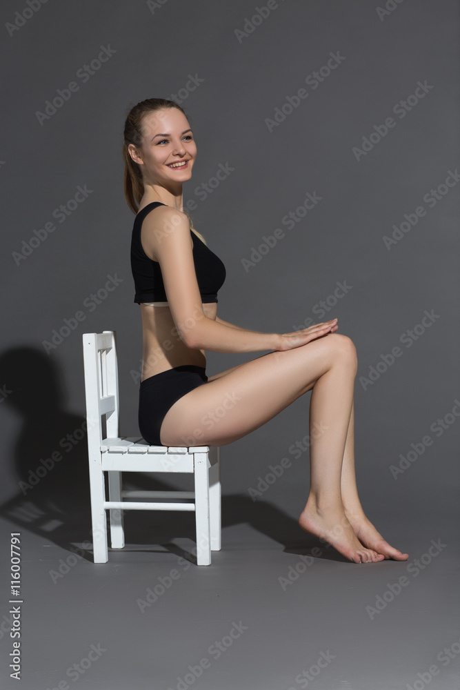 Art of seduction. Side view young woman in black posing while sitting on the chair against gray background