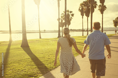 Middle aged couple walking together holding hands along the beach boardwalk. Abstract image of Building relationships and marital bonds. Early evening sunset and warm summer tone