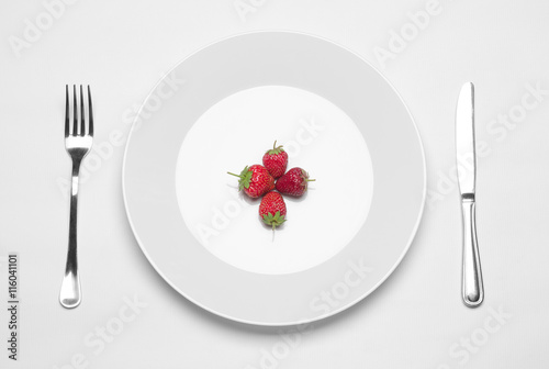 Healthy eating in the restaurant and diet Topic: white plate with three strawberries and a metal knife and fork lying on a white table in the studio isolated top view