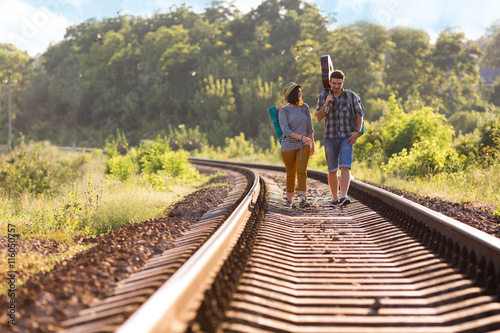 Two young People Man and Girl in casual Travel Clothing with Backpacks and Guitar walking along Railroad with back light Sun and Forest on Background