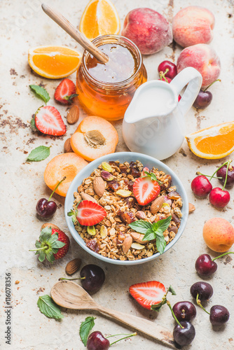 Healthy breakfast ingredients. Oat granola in bowl with nuts, strawberry and mint, milk in pitcher, honey in glass jar, fresh fruits, berries on light concrete background