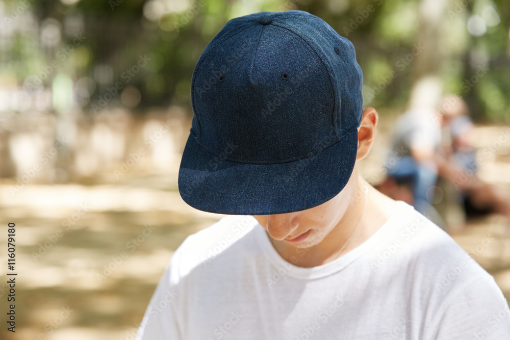 Close up view of schoolboy wearing dark blue blank snapback with copy space  for your advertising content. Depressed teenager hiding his face under  denim cap, looking down. Selective focus Stock Photo