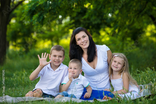 Happy mother, two sons and daughter dressed in white shirts are sitting on the grass in the park