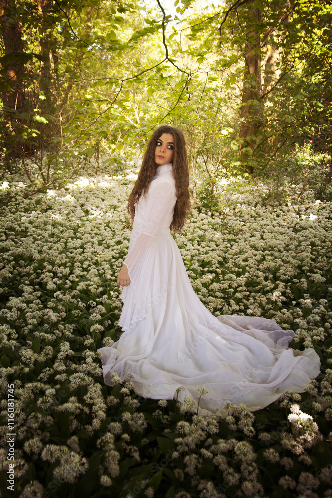 Beautiful woman in long white dress standing in a forest on a carpet of flowers