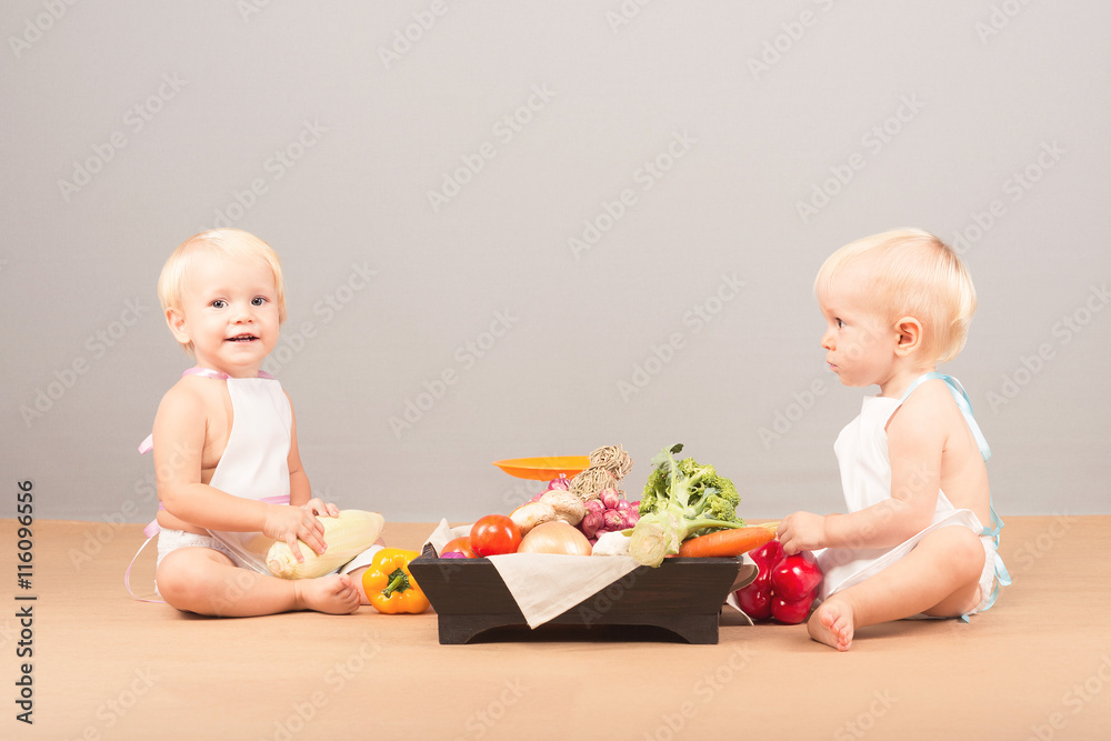 Two little boys in aprons sit next to the weights and vegetables. Basket with mushrooms, carrots, tomatoes, broccoli, red and yellow peppers, onions, corn.