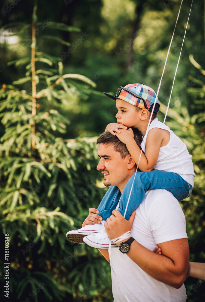 Father and son having fun in the park on a sunny day. Son on father shoulders keeps hands on the head of his father.