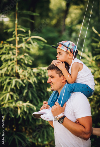 Obraz na płótnie Father and son having fun in the park on a sunny day. Son on father shoulders keeps hands on the head of his father. w salonie