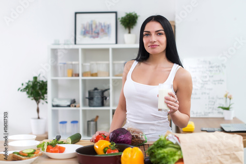 Young woman standing next to kitchen table with fresh vegetables drinking milk in the morning.