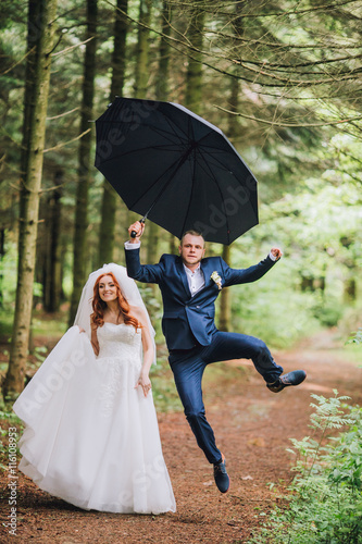 Beautiful bride and groom walking under umbrella at park. Bride with red hair. Pine forest. Newlyweds walking.