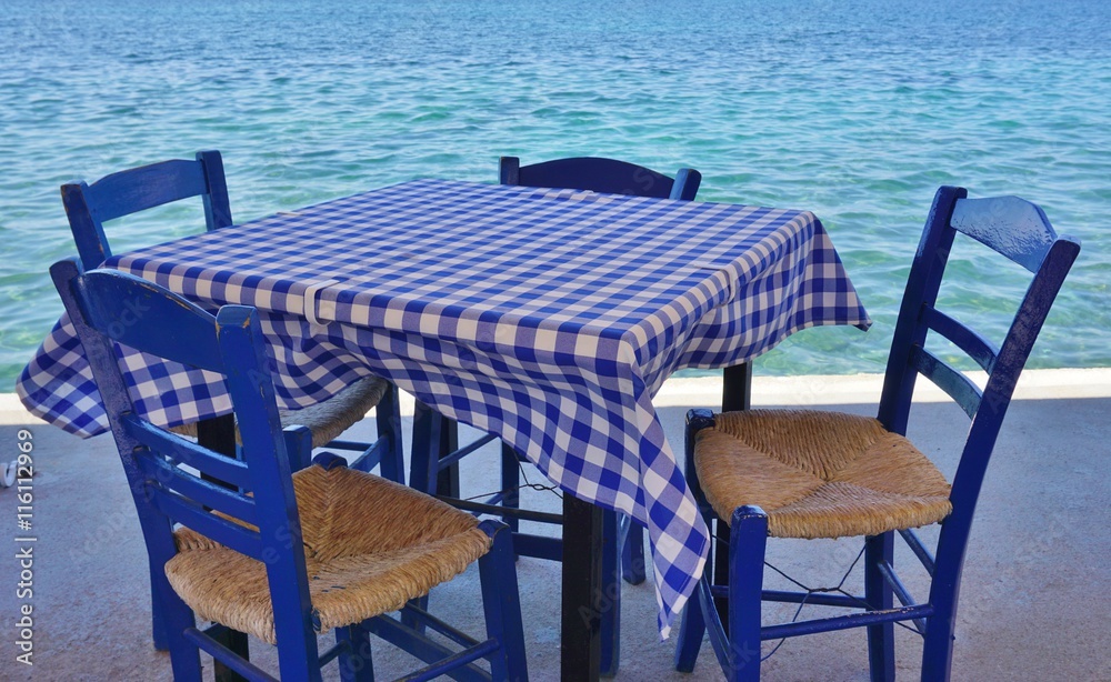 Table with a blue checkered tablecloth and blue chairs by the sea in