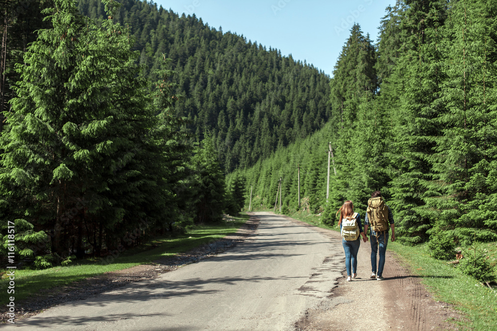 The couple go with travel backpacks on the edge of the road near a pine forest. Active holidays in the mountains