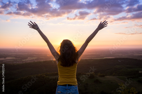 une jeune femme de dos , levant les bras face à un paysage sous le coucher de soleil