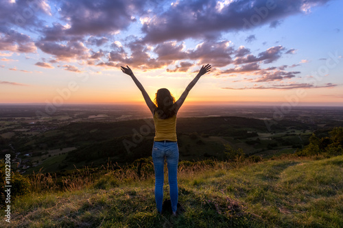 une jeune femme de dos , levant les bras face à un coucher de soleil, en haut d'une montagne