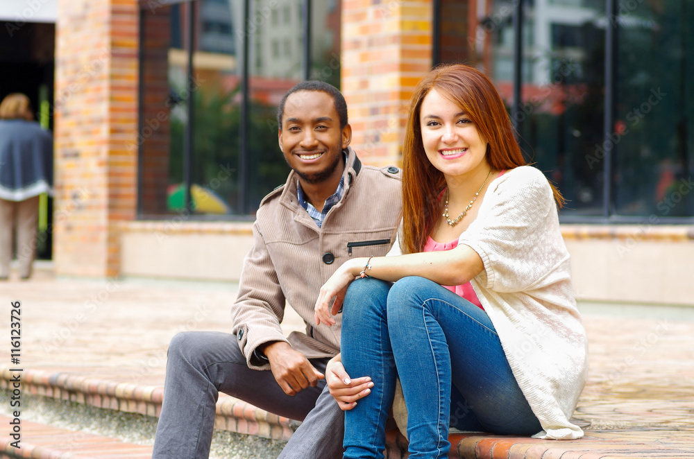 Interracial happy charming couple sitting on steps in front of building interacting and smiling for camera