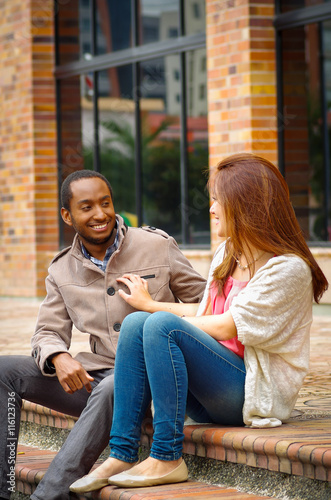 Interracial happy charming couple sitting on steps in front of building interacting and smiling for camera
