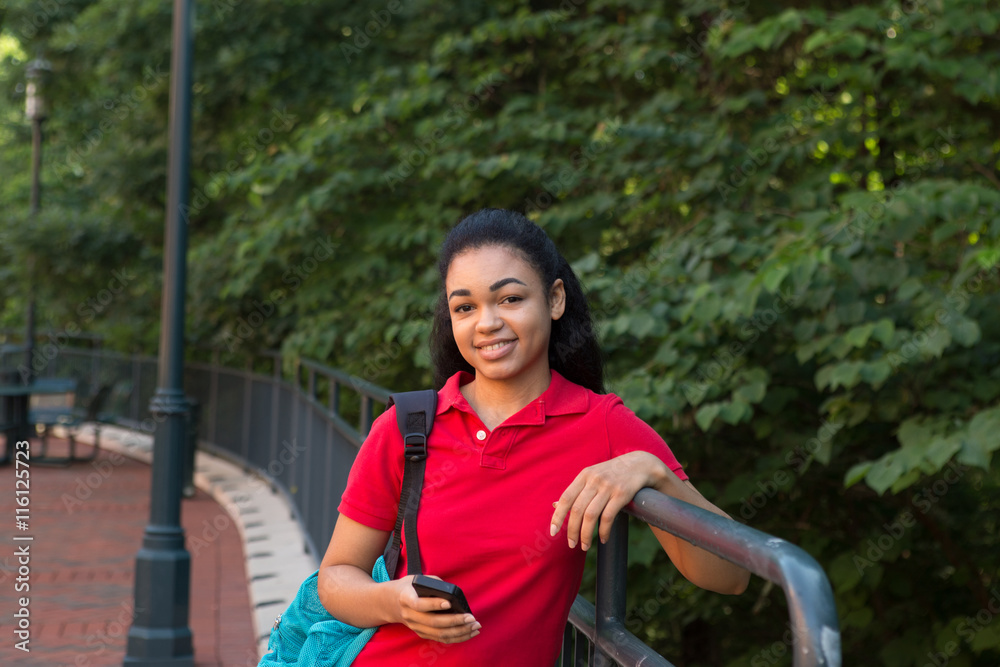 African american college student with a backpack standing by a fence holding her cell phone African american college student with a backpack standing by a fence holding her cell phone