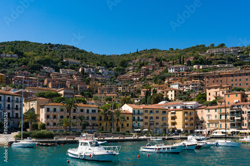 Porto Santo Stefano buildings and bay with fishing boats and yachts on a sunny summer day. Mediterranean Italian resort village of Tyrrhenian Sea with luxury property on the background © Olga K