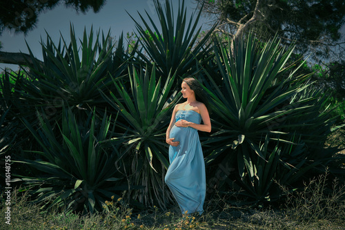 Expectant mother in a blue dress on a background of cacti. Pregnant posing against a background of green thorns.