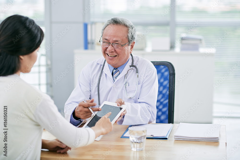 Mature general practitioner showing something on tablet computer to his client
