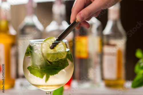 Tongs hold slice of lime. Mint leaves in beverage. Sweet syrup and sour juice. Chilled hugo cocktail.