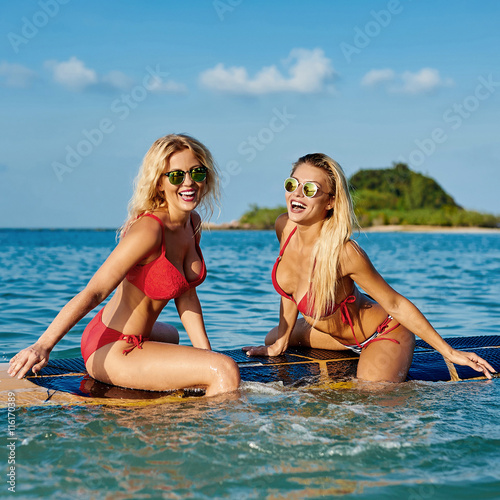 Two pretty slim tanned women having fun with surfboard on the wa