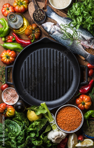 Raw uncooked seabass fish, vegetables, grains, herbs, spices and olive oil on chopping board, iron grilling pan in center with copy space, top view