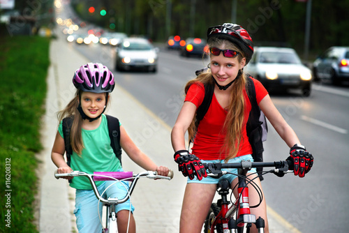 Bikes bicyclist girl. Girls wearing bicycle helmet and glasses with rucksack ciclyng bicycle. Girls children cycling on yellow bike lane at city street. There are cars on road. photo