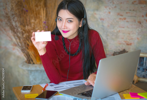 Beautiful freelancer female posing and working on line with headset with mic and looking at camera in a little office desktop or home,showing blank name card photo