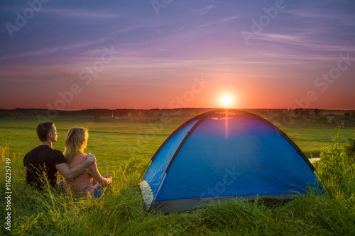 The couple sit near camping tent by sunset (sunrise) background. Wide angle