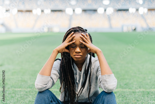 Tired pretty african american young woman sitting with legs crossed and having headache, stadium at background photo