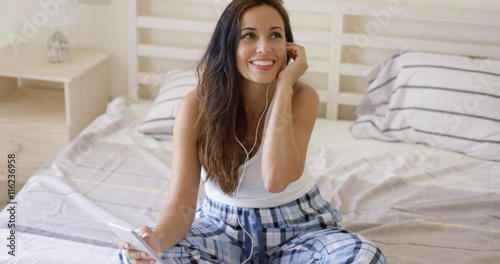 Laughing woman in blue plaid pajamas sitting in large bed listening with ear buds to something interesting on digital device