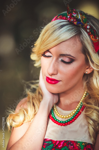 Portrait of young beautiful blonde woman wearing a long red elegant dress posing in a green meadow. Fashionable sexy attractive girl with blue eyes in field. Gorgeous fair hair female, outdoor shot.