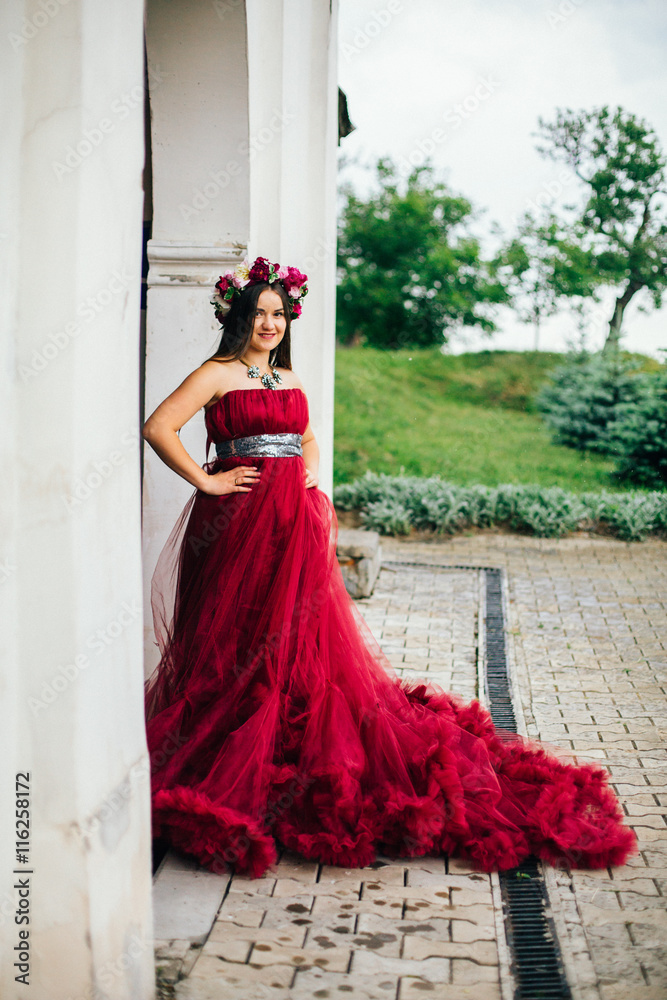 Fototapeta premium Woman in red cloud dress running near old stone castle wall and door. Girl with red roses and peonies wreath.
