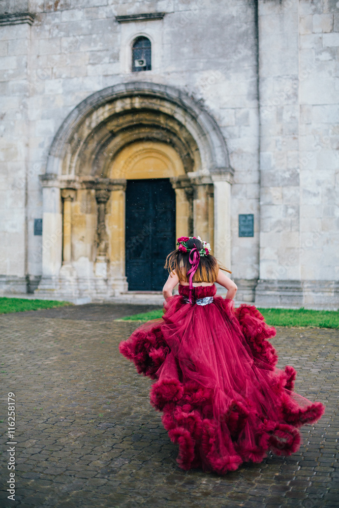 Woman in red cloud dress running near old stone castle wall and door. Girl with red roses and peonies wreath. 