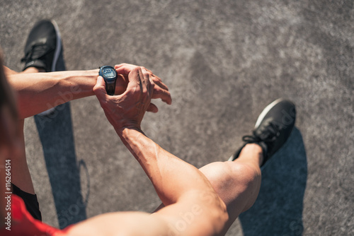 Fototapeta Naklejka Na Ścianę i Meble -  Fitness man resting during outdoor cross training workout reseting watch counter. Fit fitness sport model sitting on a bench outside before running.