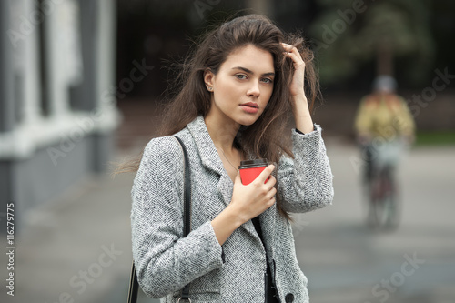 Lifestyle fashion portrait of beautiful young brunette woman in grey coat with coffee cup posing on street cloudy day
