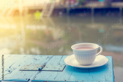 Cup of hot coffee on old wooden table and view of sunrise in the river background.Vintage tone with copy space.