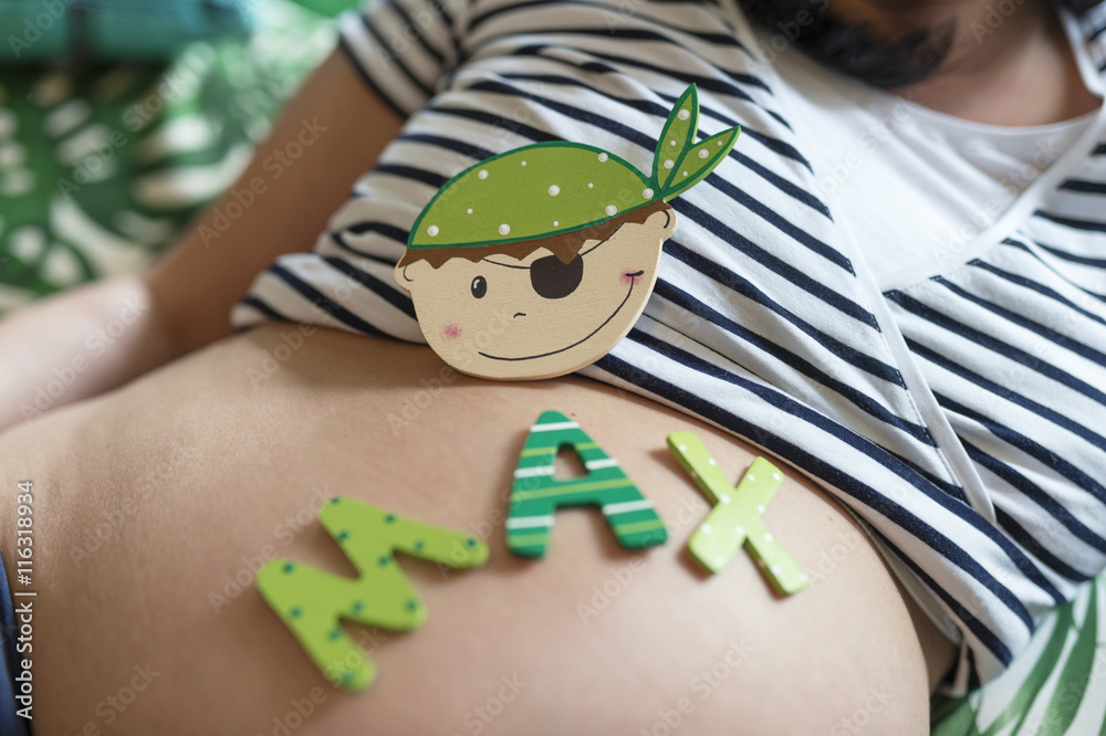 Obraz premium Auf dem Babybauch einer Schwangeren liegen ein aus Holz nachgebildetes Gesicht eines kleinen Jungen und Buchstaben zum Namen Max.