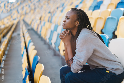 Upset african american young womanin rows of empty seats at stadium with her chin resting on hands and a glum expression