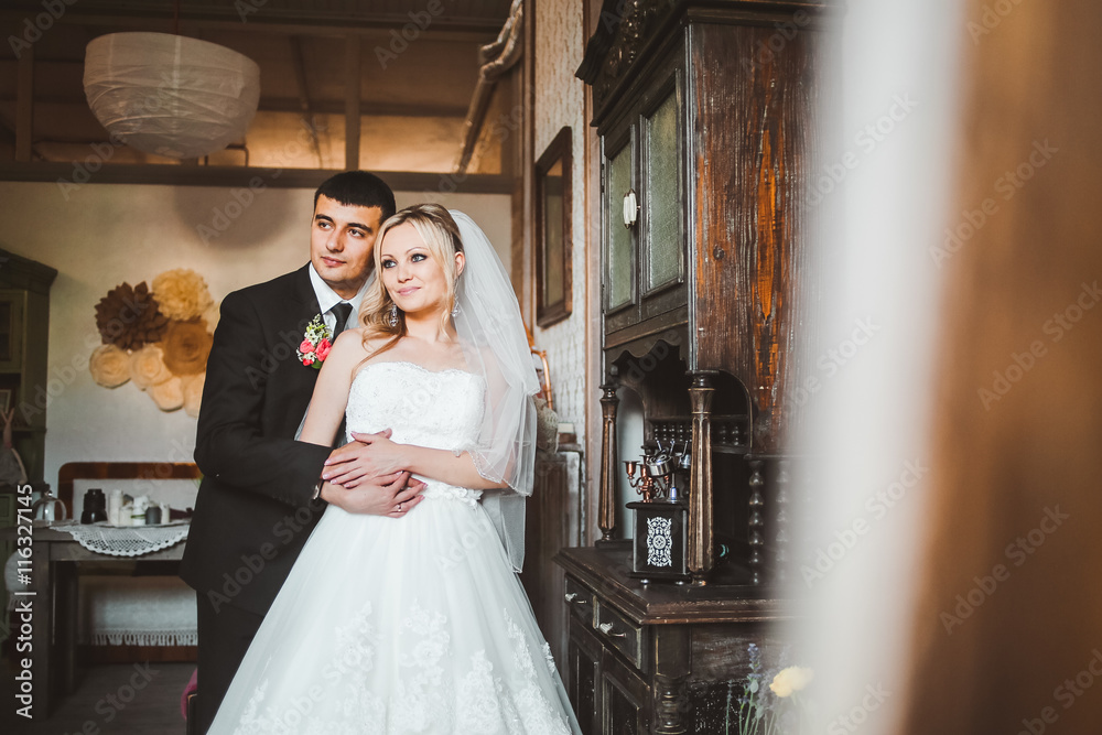 Groom hugs a pretty bride standing in the old-fashioned room. Groom holds bride's waist tender posing between old wooden mirrors