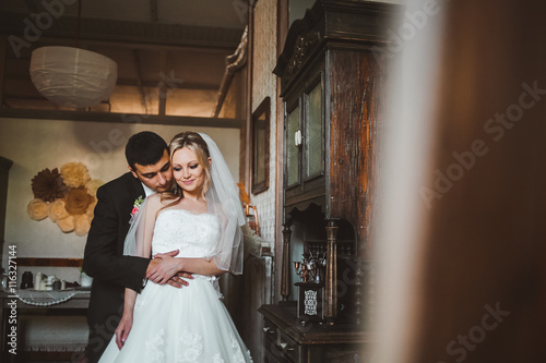 Groom hugs a pretty bride standing in the old-fashioned room. Groom holds bride s waist tender posing between old wooden mirrors
