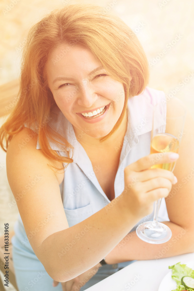 Red-haired woman in blue dress sitting in restaurant or cafe. Closeup toned image of beautiful woman holding glass of white wine and smiling to camera. Red-haired woman in blue dress sitting in restaurant or cafe. Closeup toned image of beautiful woman holding glass of white wine and smiling to camera.
