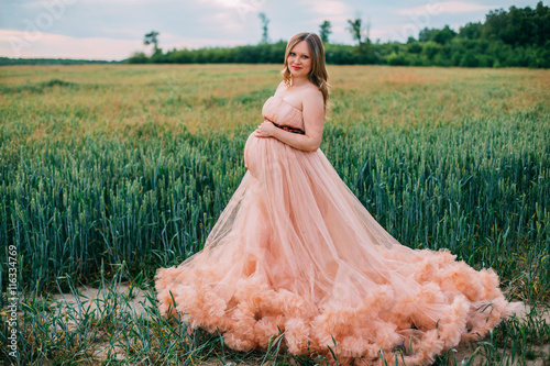 Obraz na płótnie Young happy pregnant woman in peach color cloud dress relaxing and enjoying life in nature. Outdoors on the nature. Pretty pregnant woman holding her tummy. Cloudy dress. Blues sky. Wheat green field. w salonie
