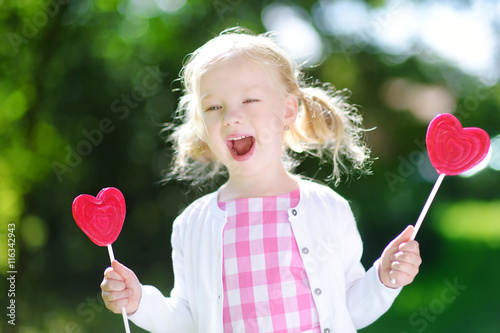 Cute little girl eating huge heart-shaped lollipops outdoors